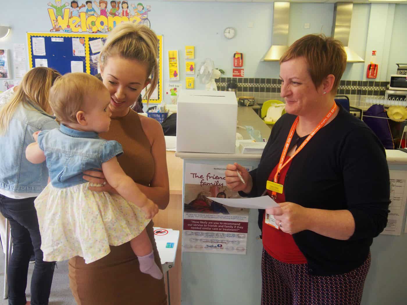 A woman with blonde hair holding a baby, looking at an older woman with short red hair who is writing something on a piece of paper. They're stood in a pre-school, with the kitchen visible in the background and on the right.