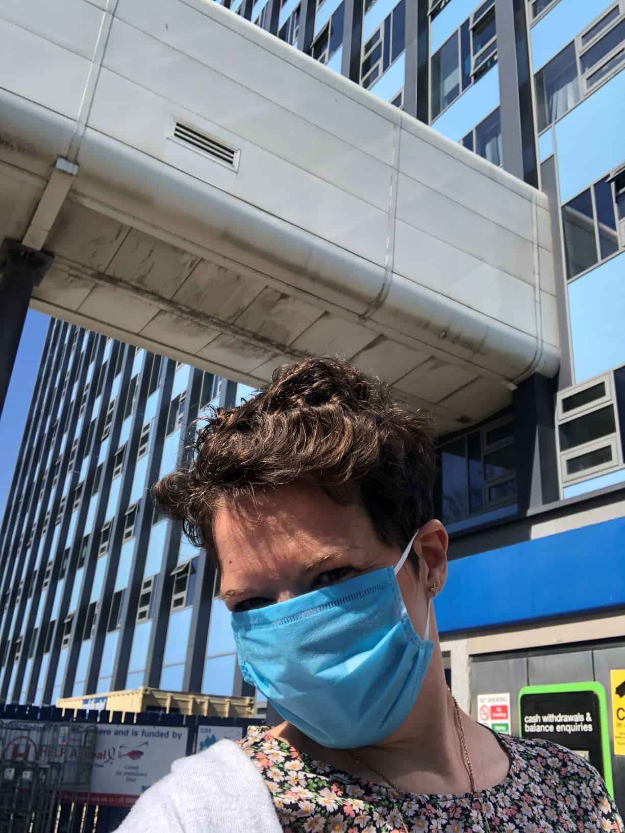 This photo is of Lizzie stood outside Hull University Teaching Hospitals Trust main tower block. There are windows surrounded by blue cladding and Lizzie is wearing a blue facemask, floral t-shirt and grey cardigan. She has short brown hair.