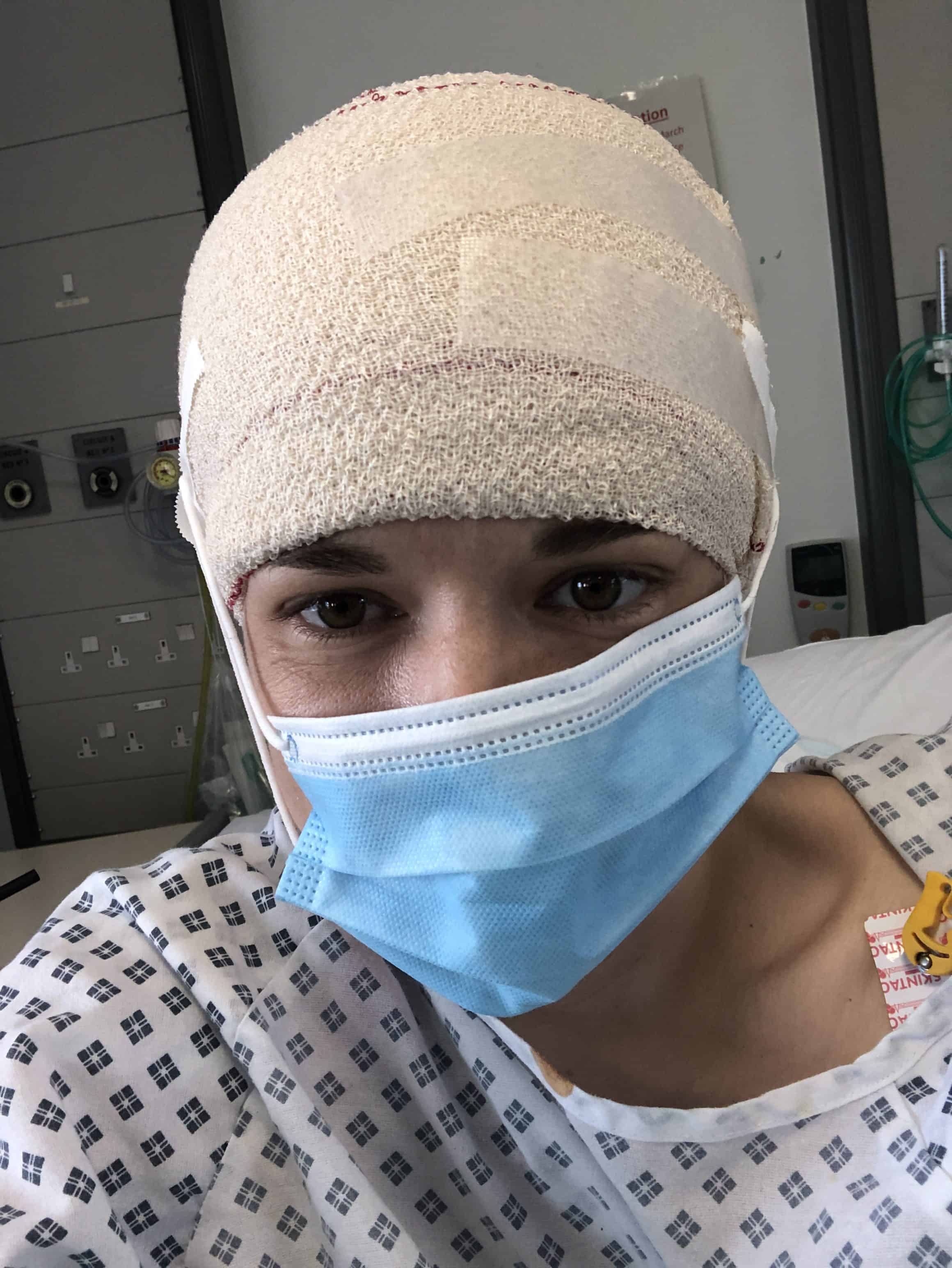 This is a photo of Lizzie who is living with brain cancer during the Covid-19 pandemic. Lizzie is on a hospital bed and the image is of her shoulders and head. She is wearing a hospital gown, blue face mask and has a bandage around the top of her head. She is looking at the camera.
