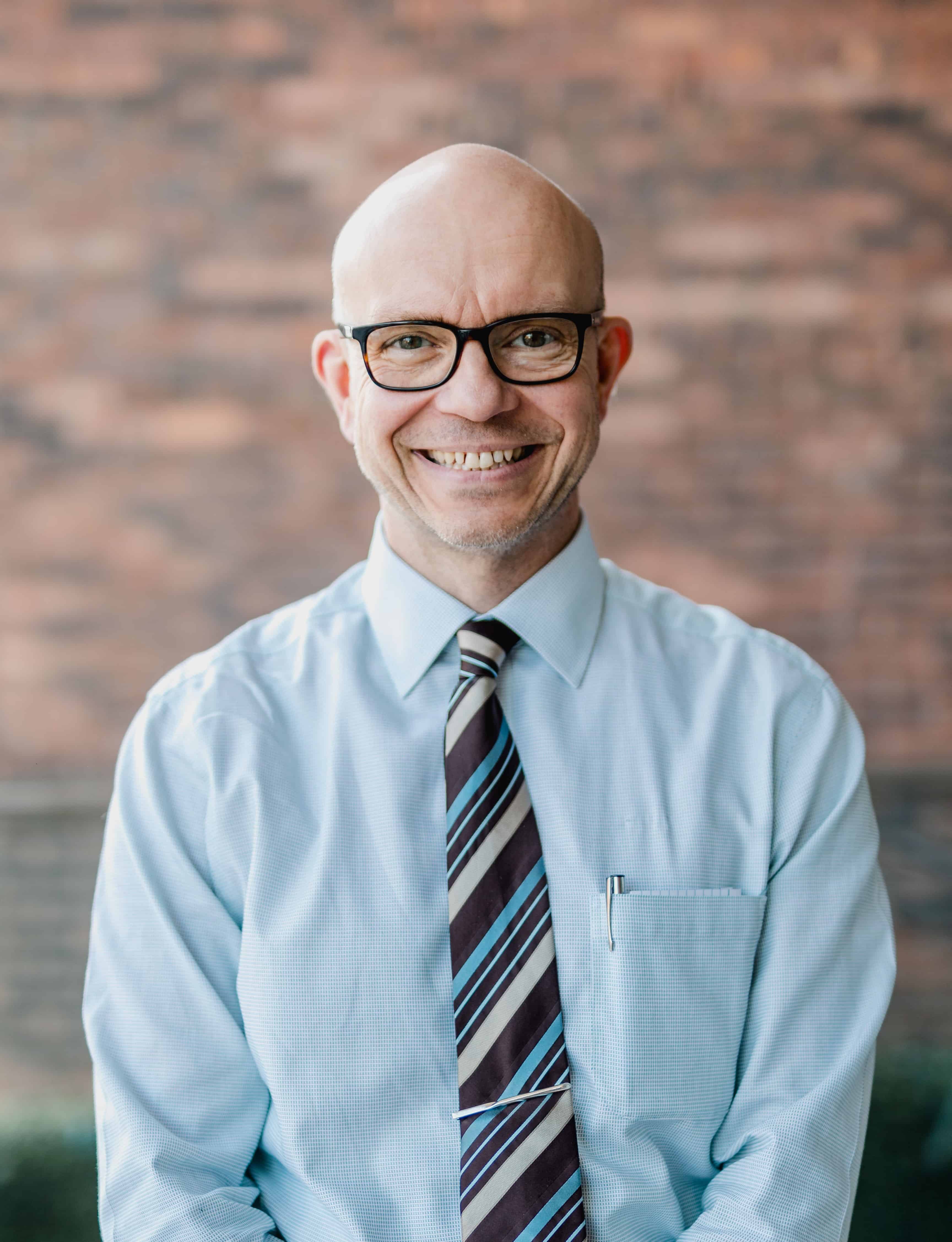 Image of Dr Dan Cottingham who is the GP Lead for Cancer at Humber, Coast and Vale Cancer Alliance. The image shows Dan standing in front of a brick wall, wearing a shirt and tie. He is wearing glasses and is smiling.