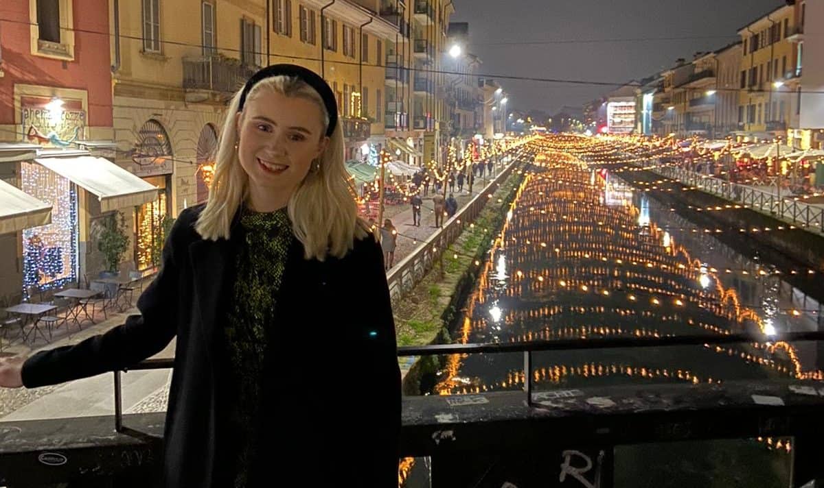 Beckie, a girl in her mid-20s, standing in front of a canal in Prague, smiling at the camera. It's night time and the canal is lit up.