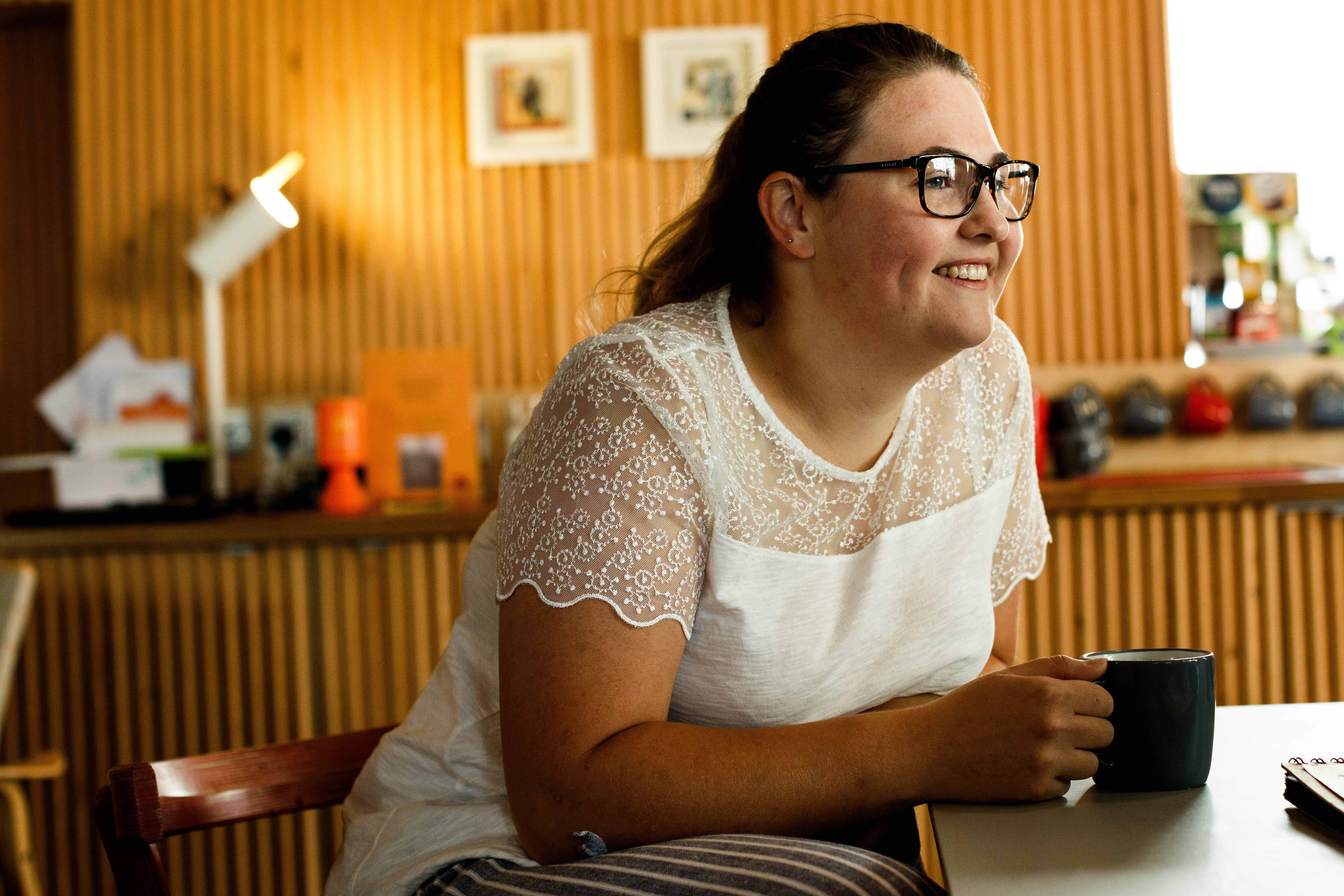 Picture of a woman sat at a table. The woman is leaning on the table and holding a mug. She is wearing a white top and her brown her is in a ponytail. The lady is wearing glasses and smiling.