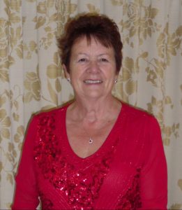 Christine, a pancreatic cancer survivor, looks into the camera and smiles. She is wearing a red sparkly long-sleeved top and has brown cropped hair.