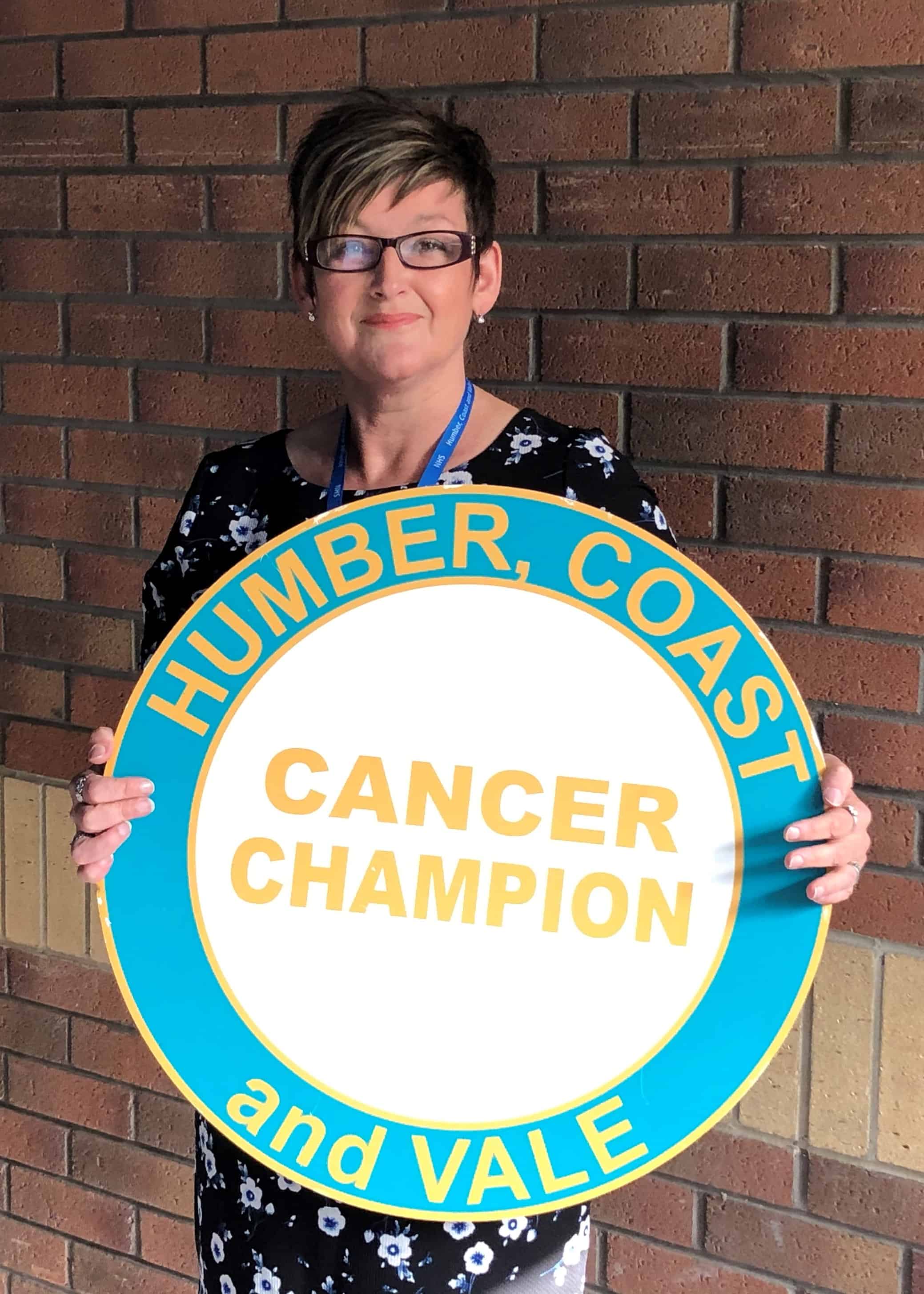 Photo of Sarah Patten stood in front of a brick wall. Sarah has short dark hair and is looking at the camera smiling. She is holding a photo prop which is of an enlarged Cancer Champion logo.