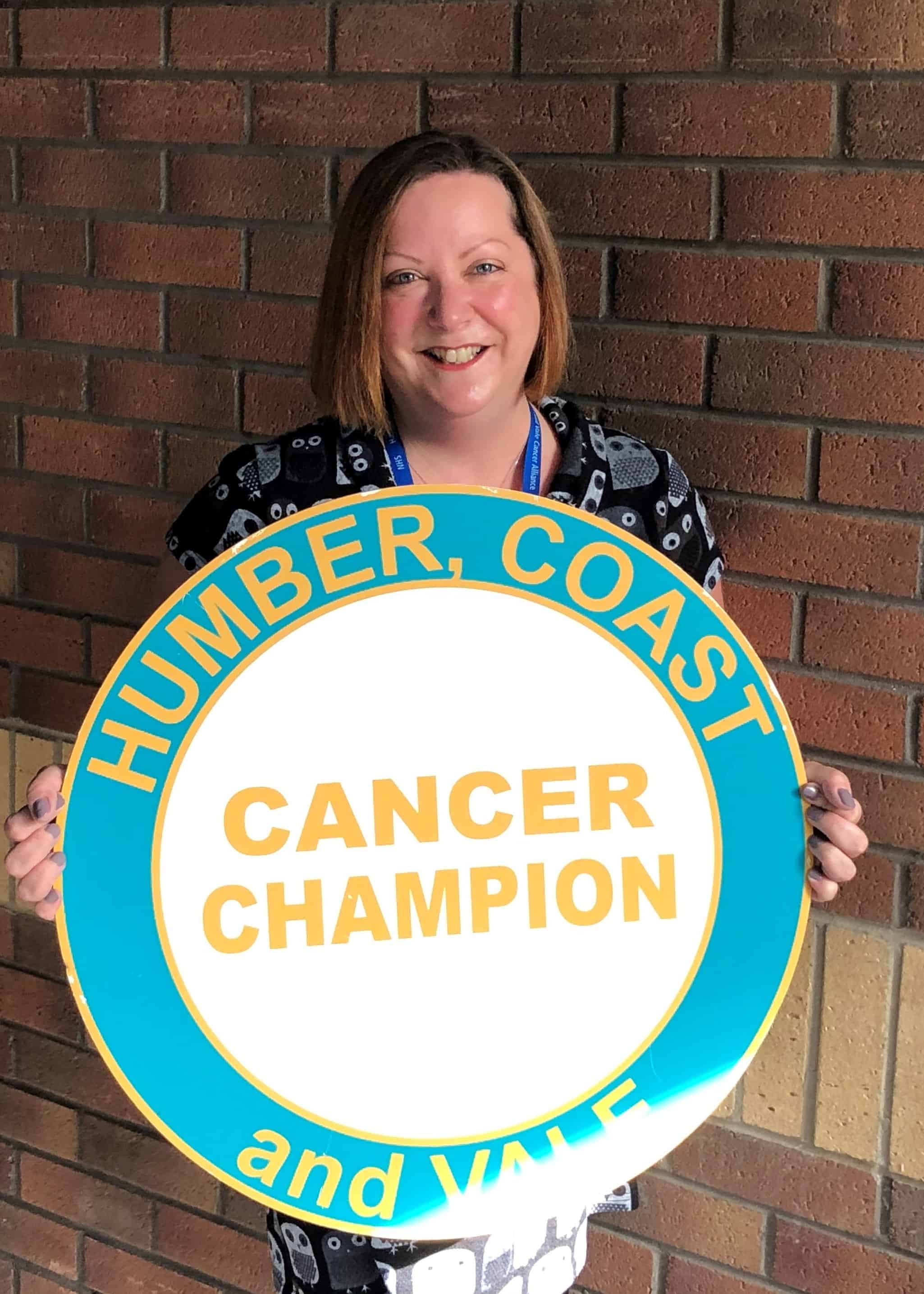 Photo of Zoe Bounds standing in front of a brick wall. Zoe is holding a photo prop of the Cancer Champion logo. Zoe is smiling and has shoulder length, dark, hair.