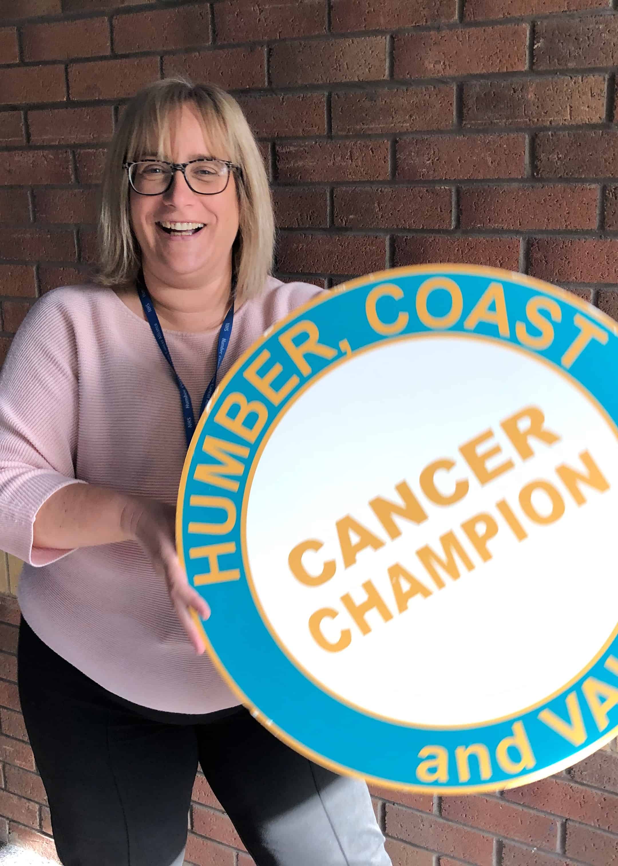 Photo of Emma Lewin, volunteer co-ordinator is stood in front of a brick wall holdig a photo prop of the Cancer Champions logo. Emma is looking at the camera and smiling. She has blonde shoulder length hair, a fringe and glasses. She is wearing a pink top and grey trousers.