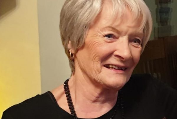 Headshot of Margaret Allbones, a cancer survivor who shared her story with us. She is smiling, looking slightly to the right of the camera, and wearing a black jumper. She has cropped blonde hair and is wearing small gold hoop earrings.