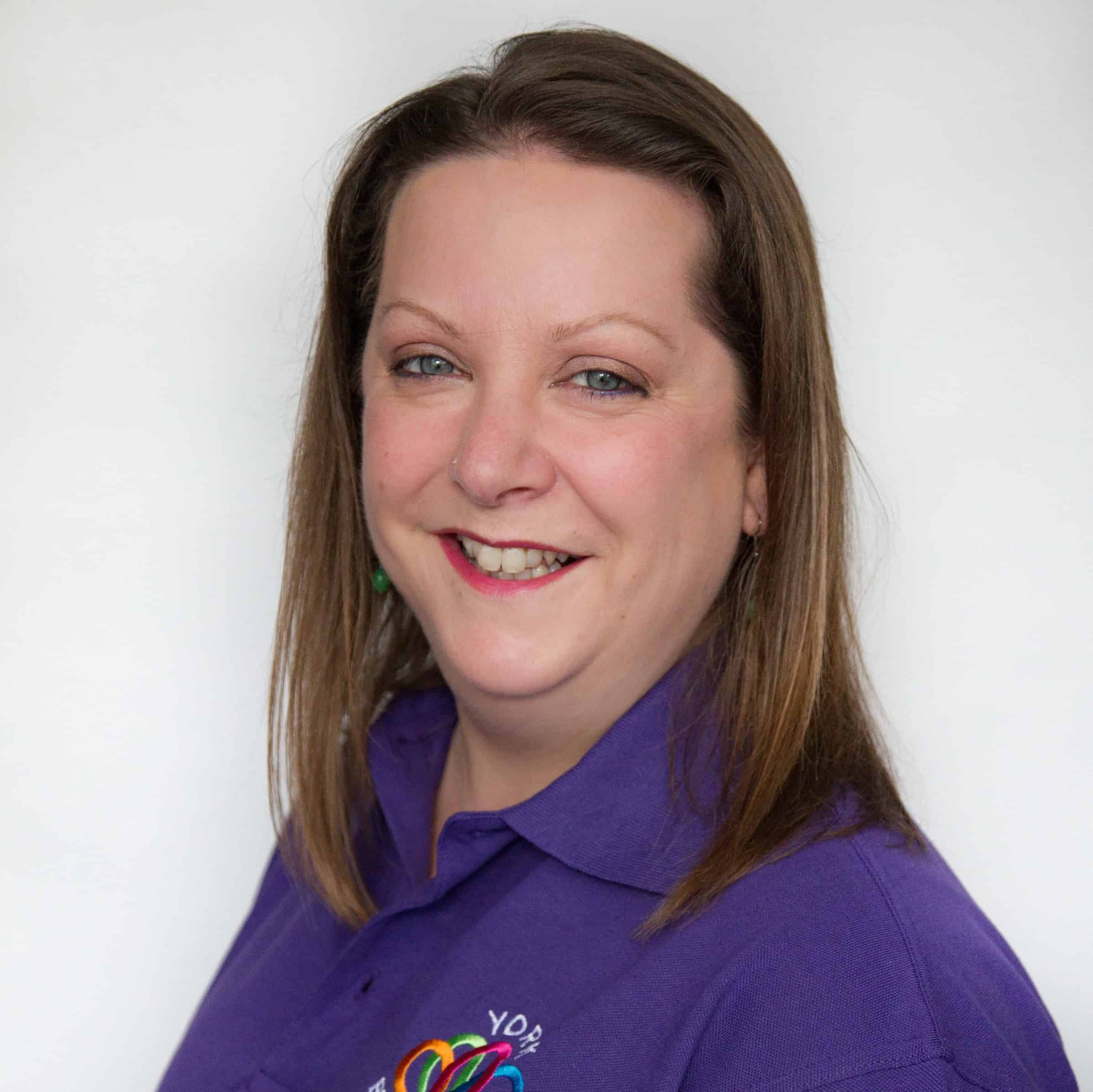 A headshot of Zoe, a woman who has survived cancer twice, smiles to the camera. She has shoulder-length brown hair and is wearing a purple 'York Breast Friends' polo shirt.