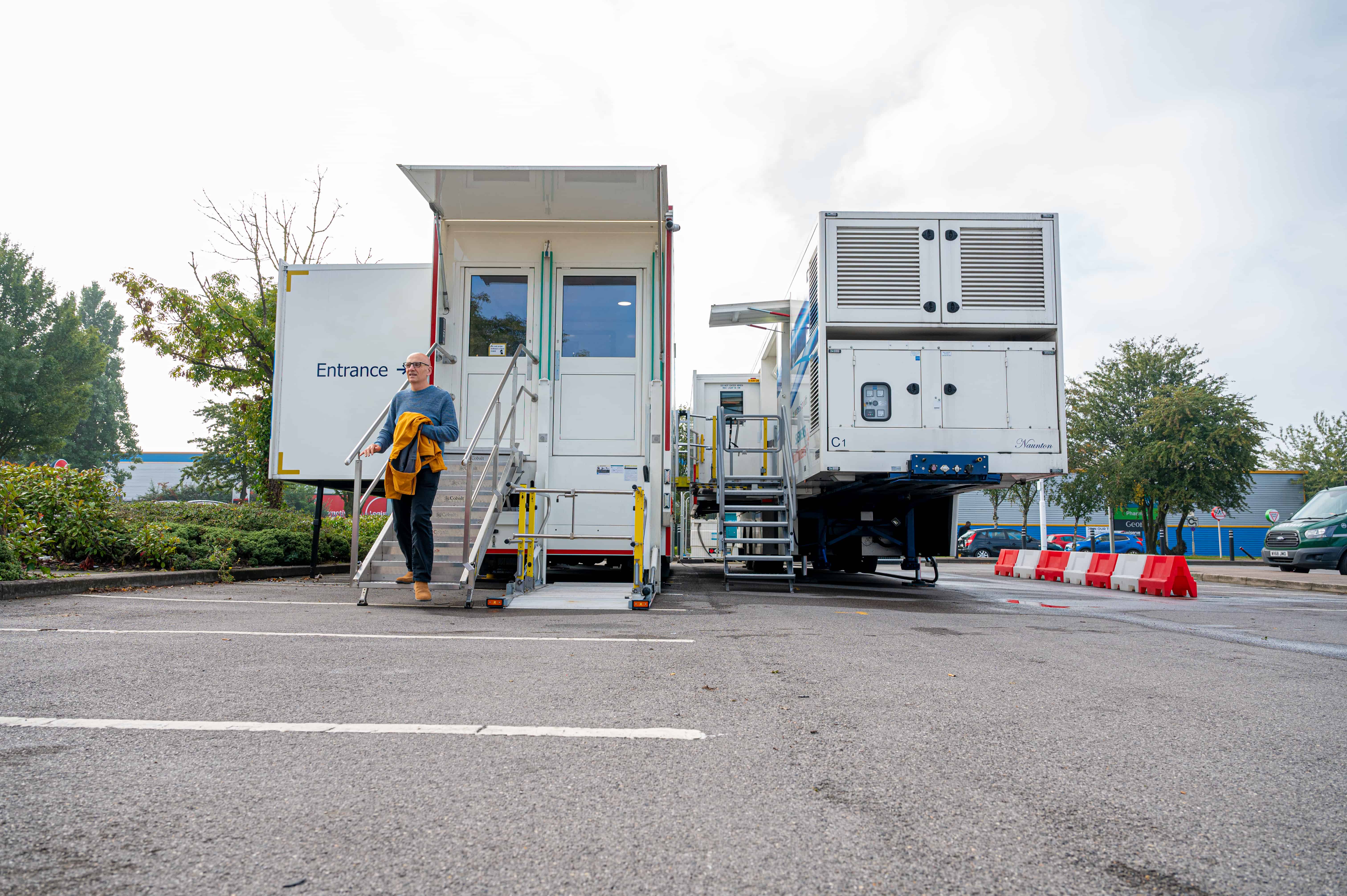 Image of a large mobile unit which is parked in a car park. A man is walking down some steps away from the unit.