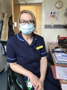 Photo of Vicky sat beside a desk. She is wearing a blue nurses uniform, glasses and a face mask. Her hair is tied back and she is looking at the camera.
