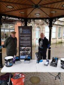 Andrew Markham and a friend fundraising for Prostate Cancer UK. They are standing outside at a table with various merchandise.