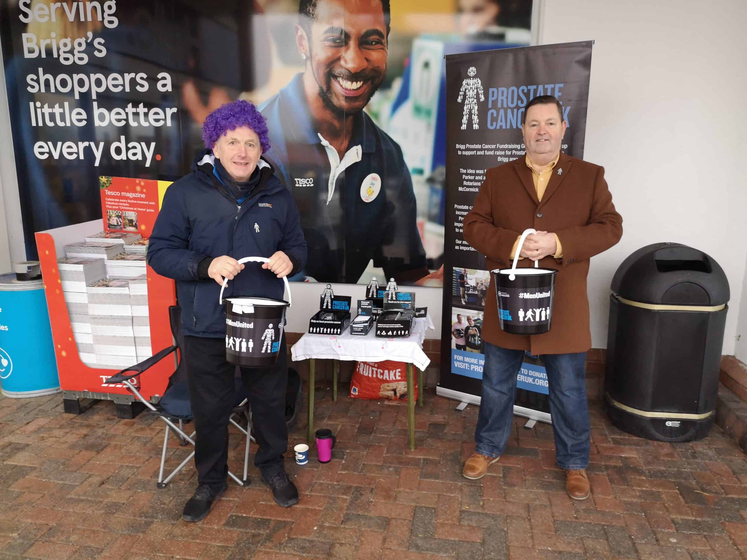 Andrew Markham and a friend raising money for Prostate Cancer UK. They are standing outside a Tesco and holding fundraising buckets. Andrew, who stands on the left, is wearing a purple wig.
