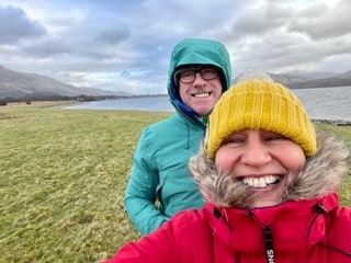 Allyson is wearing a red coat and yellow bobble hat. It is a selfie photo which also includes a man standing in the background, smiling at the camera. They are both in an open field which has a mountain and water in the background.