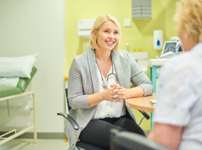 picture of a female GP in a grey jacket, black trousers and white blouse with blonde bobbed hair talking to a female patient in her consultation room. Bed to the left and sink behind with desk to the right hand side