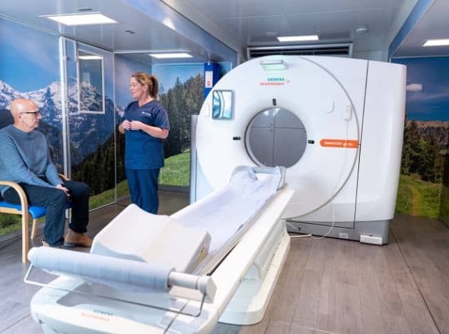 picture of a man sitting next to a CT scanner and being talked to be a female radiographer