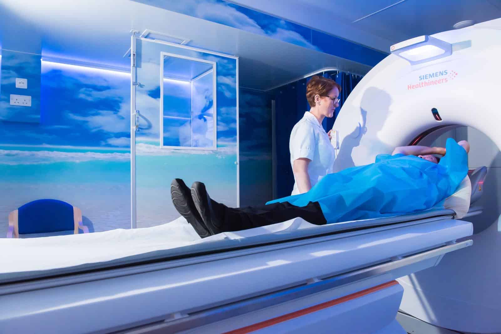 picture of a person in a blue hospital gown laying on the bed of a CT scanner with their hands above their head and a nurse next to them operating the scanner