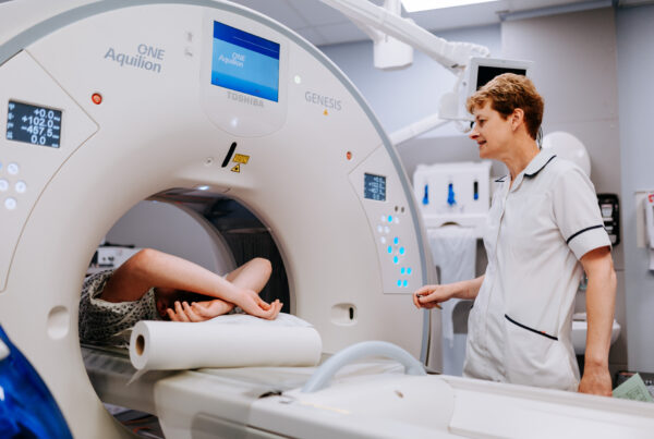 Person inside a CT scanner with a lady in a radiographer's uniform stood to the right hand side operating the scanner