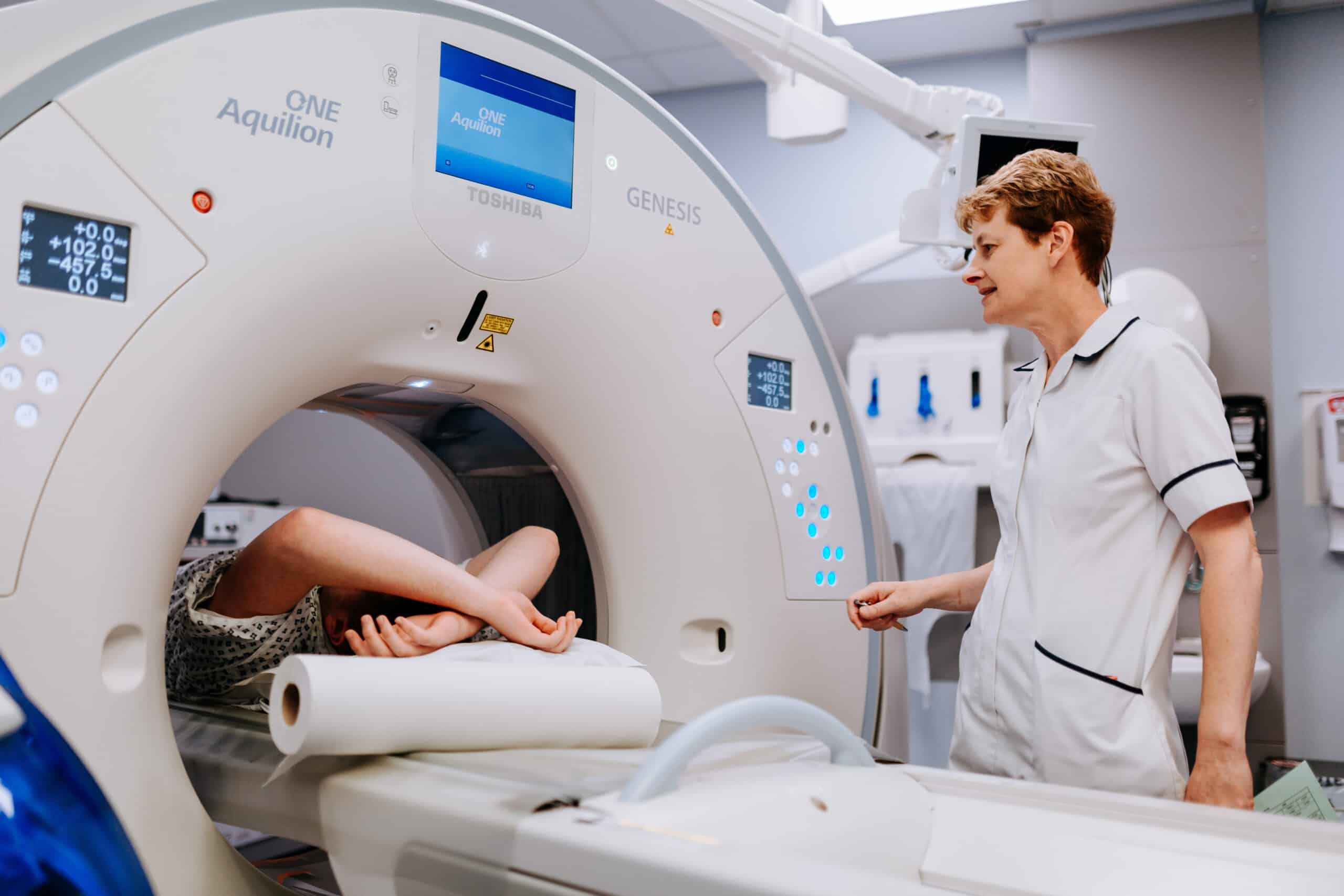 Person inside a CT scanner with a lady in a radiographer's uniform stood to the right hand side operating the scanner